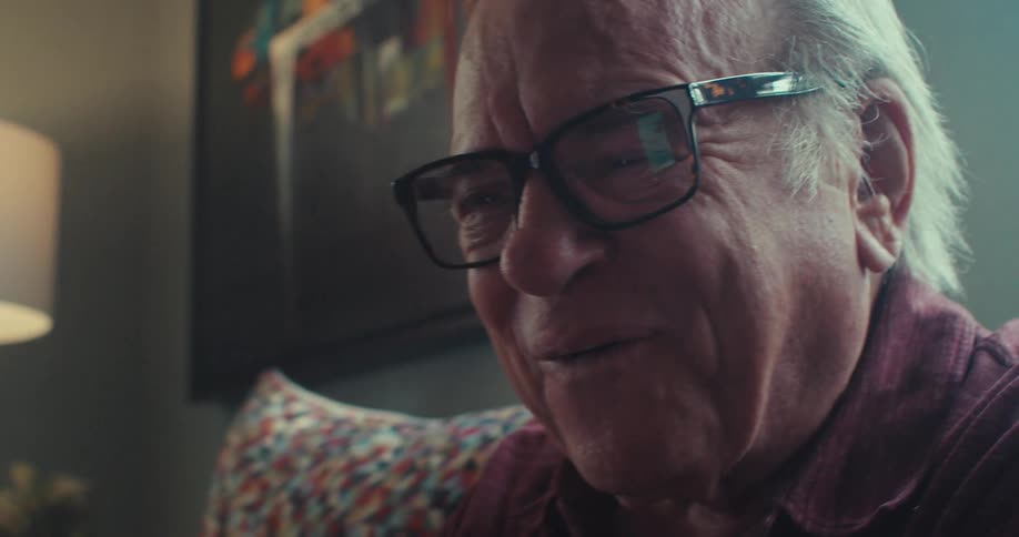 An elderly man in glasses smiles and laughs while talking on the phone in his home