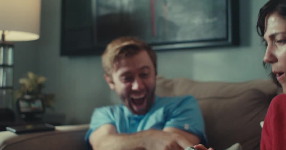 A man and a woman are sitting on the couch playing video games and the man makes a joke with the woman and starts laughing