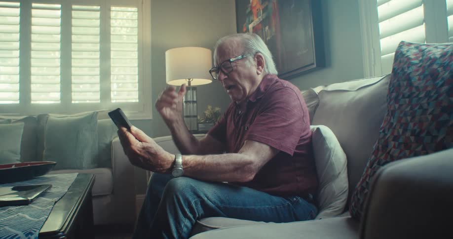 An elderly man sits alone on a couch during the day looking thoughtfully at a phone in his hand