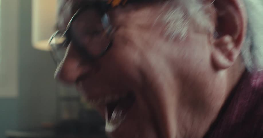 An elderly man wearing glasses is sitting on a sofa using his cell phone and is very happy while looking at it
