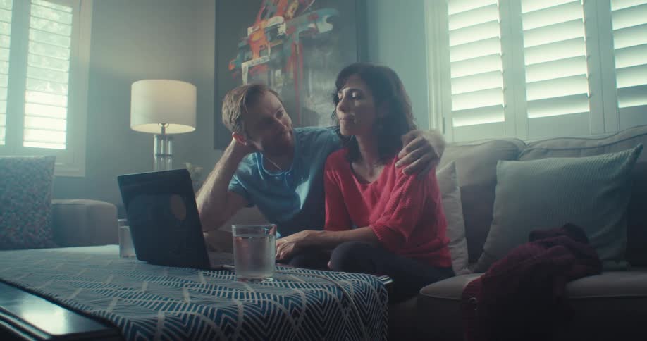 A man wraps his arm around a woman as they sit close focused on a laptop