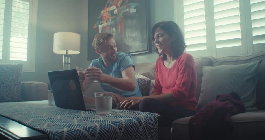 A couple smile and embrace while conversing about contents seen on a laptop screen in a living room