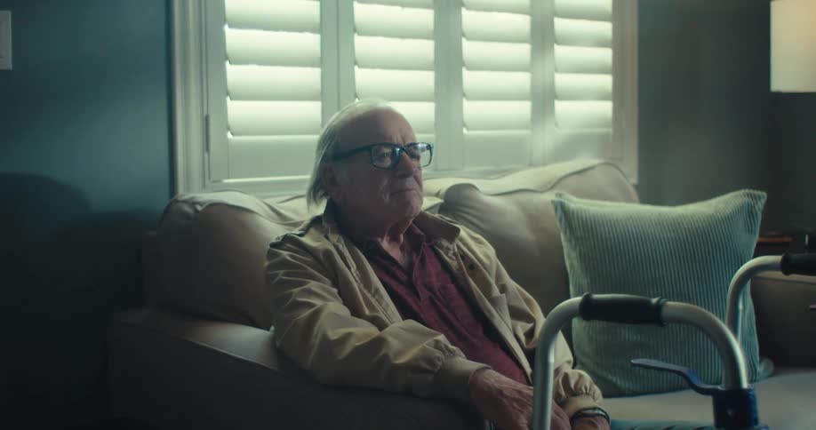 An elderly man sits by his walker gazing out a window in soft light