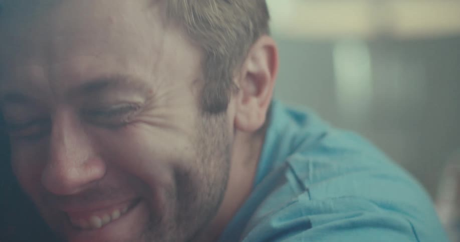 Close up of a smiling man with a beard and short hair hugging a woman sitting next to him in a sunlit room