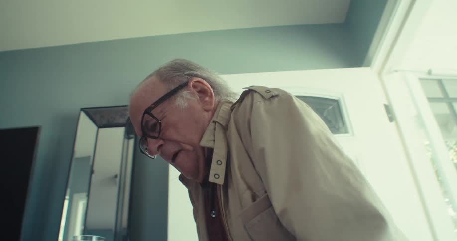 An elderly man wearing glasses walks into a house using a walking frame