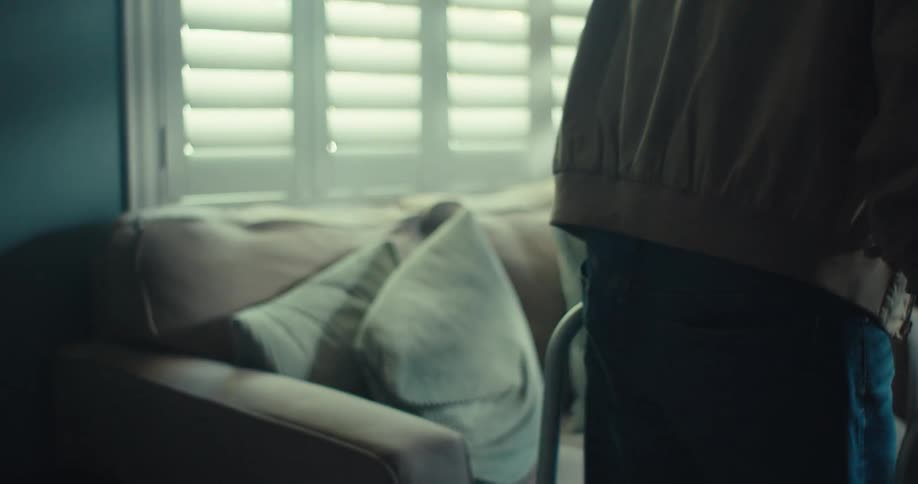 Elderly man walks towards a couch in a living room with the aid of a walker and sits