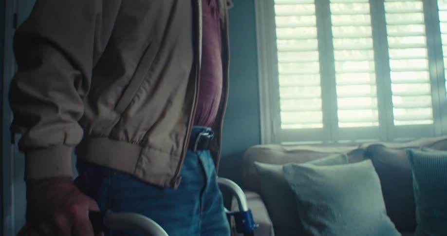 A man enters his home and walks to the couch using a walker