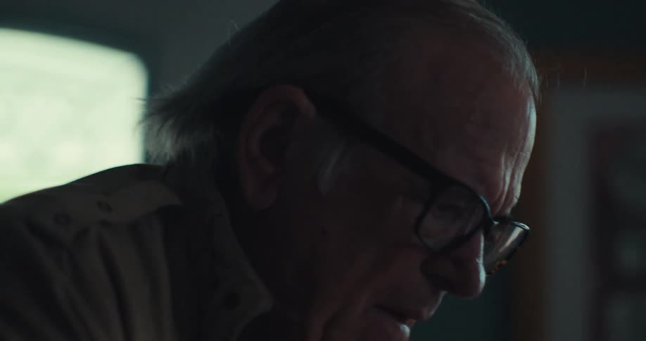 An elderly man wearing glasses walks into a house using a walking frame and then sits down on a couch