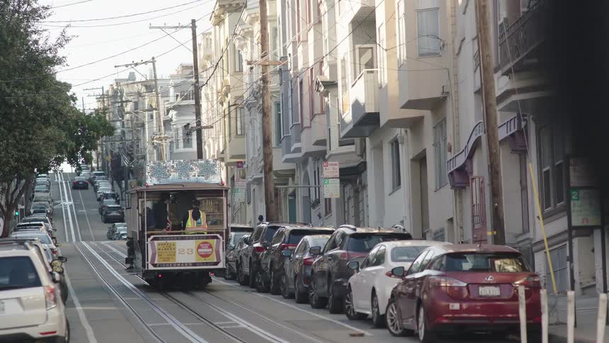 A cable car with people on board is making its way up a steep street lined with parked cars and multistory buildings