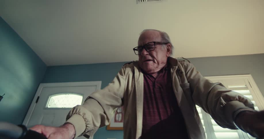 An older man in glasses a lightcolored jacket and a reddish shirt sits down carefully on a chair viewed from the perspective of his walker
