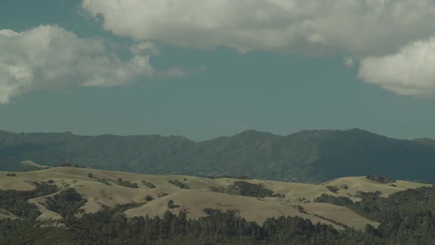 Vast rolling hills covered in dry grass and scattered trees stretch into the distance with a ridge of mountains under a partly cloudy light blue sky in the background