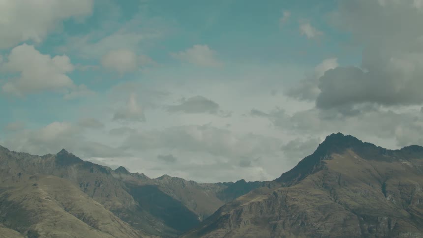 A rugged mountain range with sharp peaks and shadowed valleys stands under a blue sky scattered with white clouds