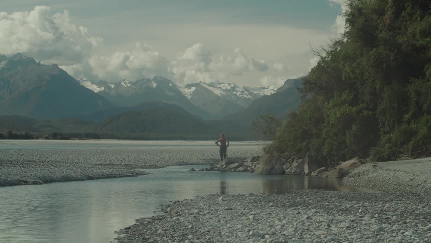 A lone person stands by a river with rocky ground and snowy mountains behind