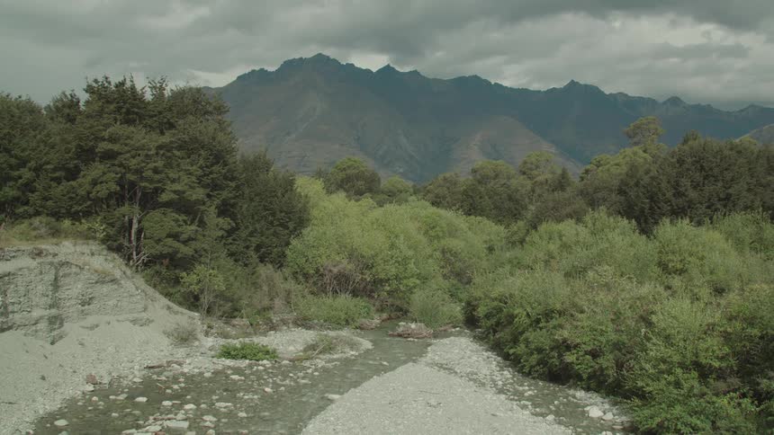 A narrow rocky river winds through a lush green landscape with dark imposing mountains under a cloudy sky in the background