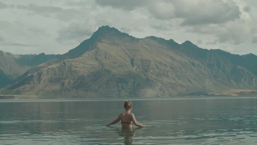 A woman in a swimsuit is standing in a calm body of water facing away from the camera with a large rugged mountain range under a cloudy sky in the background