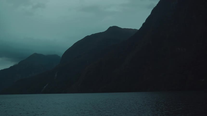 Dark moody mountains loom over a still body of water under an overcast sky