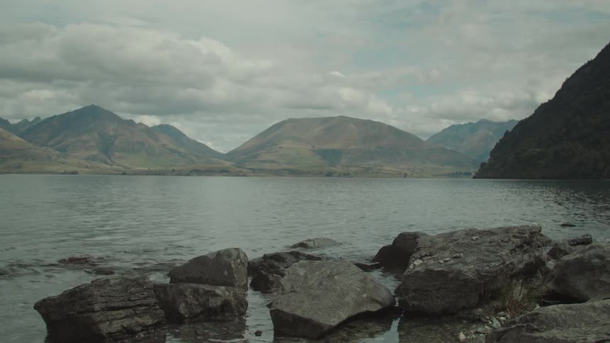 A vast lake with rocky shores is surrounded by large rugged mountains under a cloudy sky