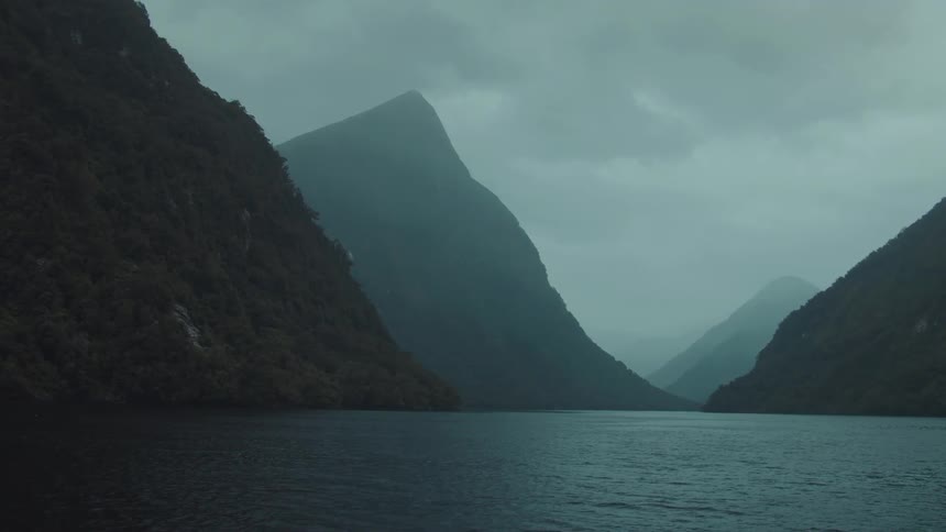 Steep treecovered mountains recede into the distance framing a dark calm fjord under a heavily overcast and moody sky