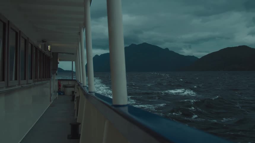 A boat sails on choppy waters with distant mountains under a cloudy sky