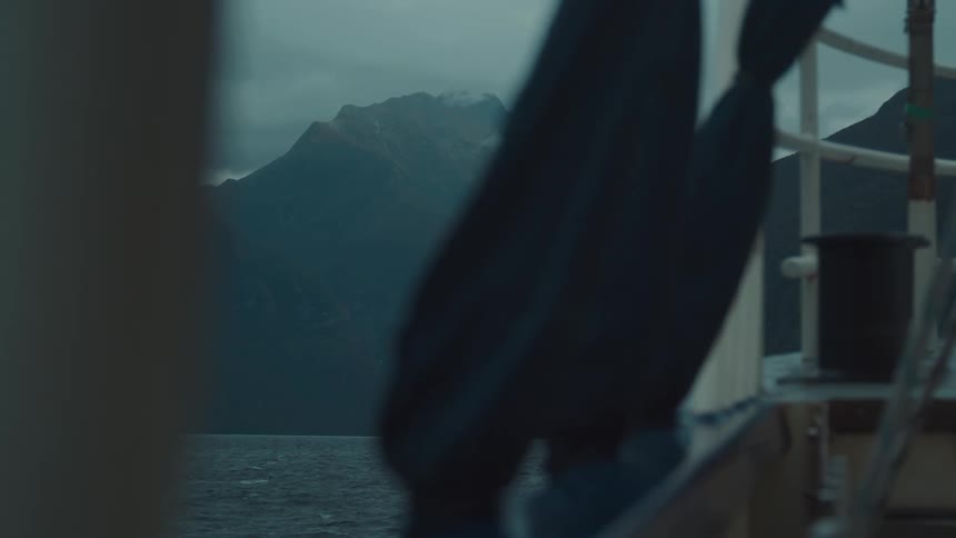 A view from a boat looks past a blurred foreground object towards a distant misttopped mountain across a fjord under an overcast sky