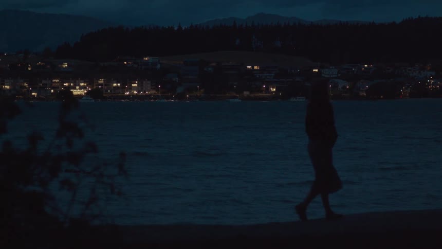 A silhouetted figure walks at night as the lights of a town twinkle across the bay