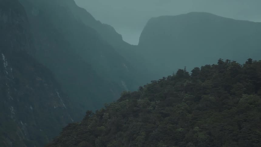 A dark mistcovered mountain range with a dense forest in the foreground stands under an overcast sky