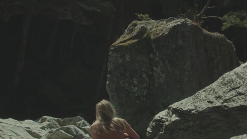 A woman with long lightcolored hair sits in front of a large grey boulder looking away from the viewer towards dark mountains