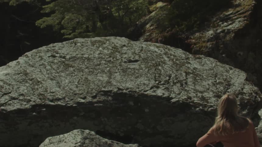 A woman sits beside a giant rock in a forested area during daylight