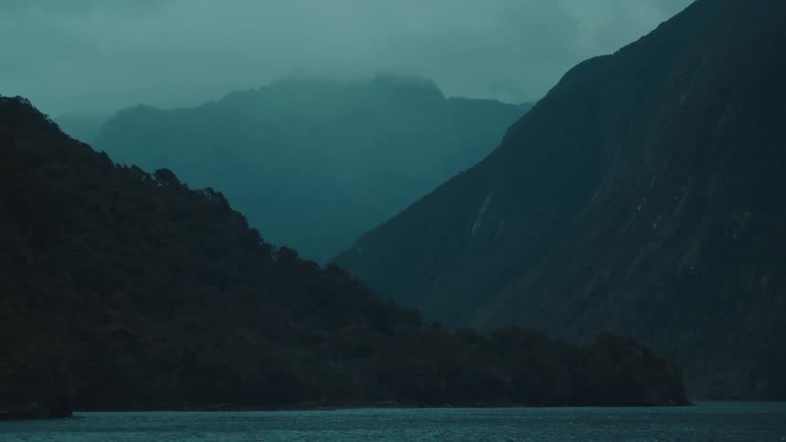 Silhouetted mountains rise steeply from a dark body of water with layers of foggy peaks visible in the distance under a moody overcast sky