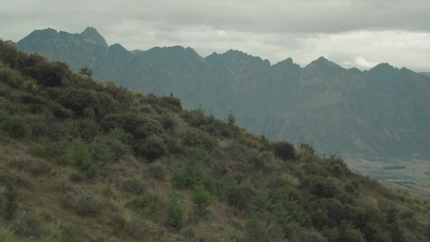 A vast landscape features a steep brushcovered hillside in the foreground with jagged mountain ranges under a cloudy sky in the background