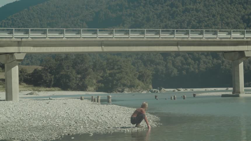 A person kneels by a calm riverbank under a bridge gently touching the water