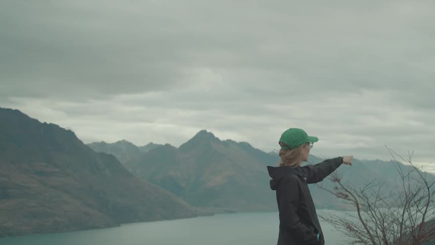Person in green cap points at mountains beside a lake under grey skies