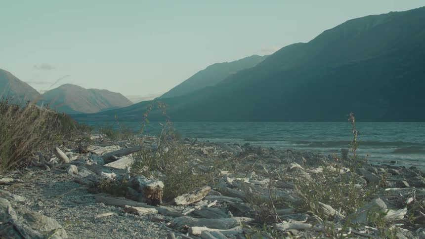 A quiet rocky lakeshore lies beneath towering mountains under a soft midday sky