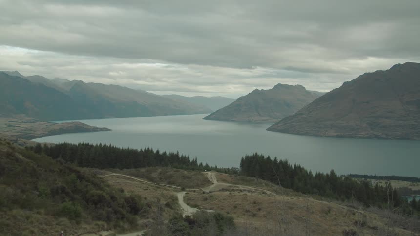 A footpath leading toward a lake surrounded by mountains under a cloudy midday sky