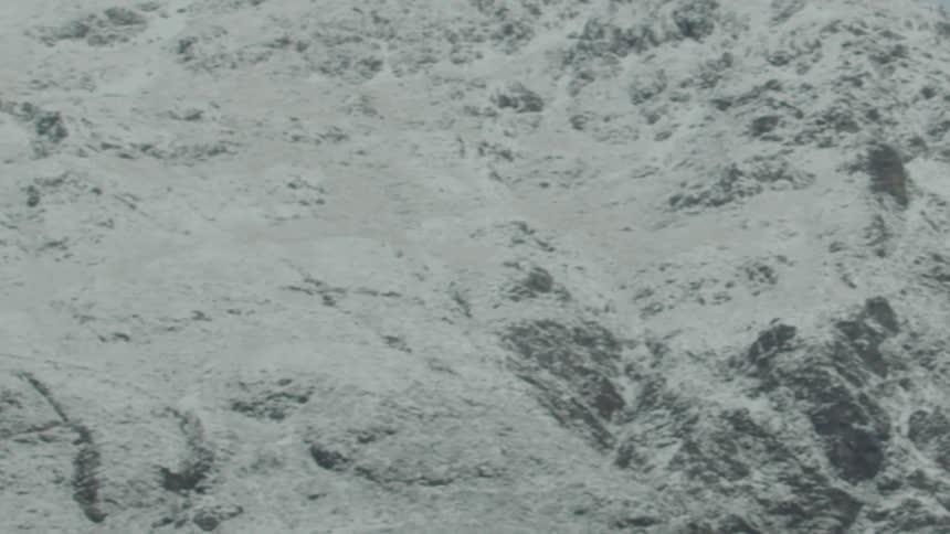 An aerial shot pans across a vast rugged mountain range entirely covered in snow and ice eventually tilting slightly towards a pale overcast sky