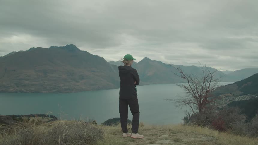 Woman stands on a hill looking at a lake surrounded by mountains on a foggy overcast day