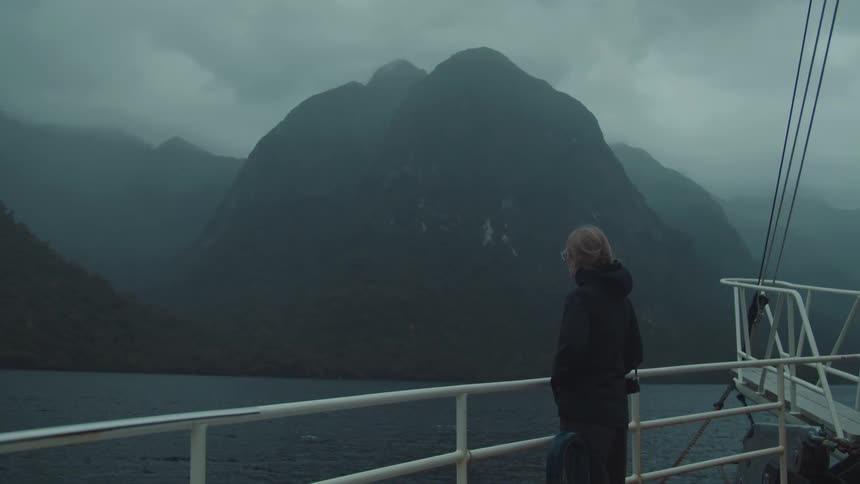 A person stands on a boat gazing at misty mountains across a large body of water