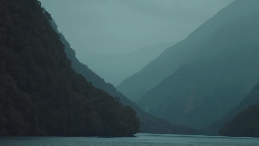 A serene view of a calm river with tall dark mountains rising steeply on either side under a misty sky