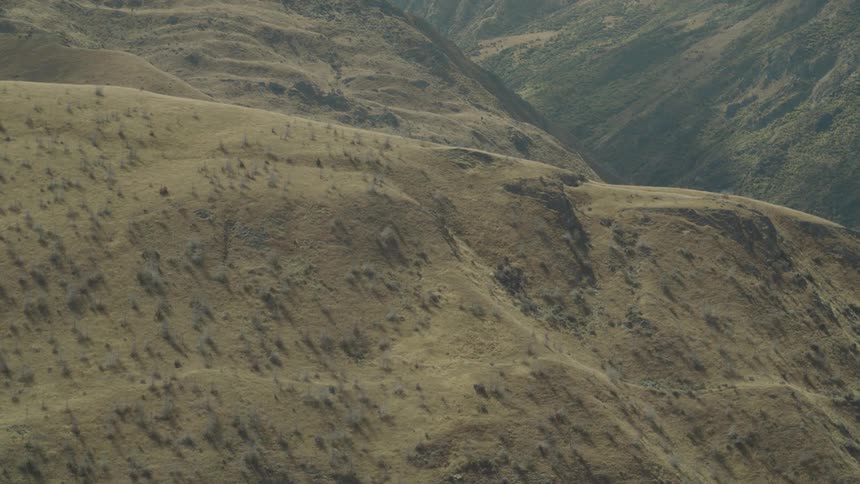 An aerial perspective shows dry rolling hills sparsely dotted with small bushes with a deeper valley visible in the background