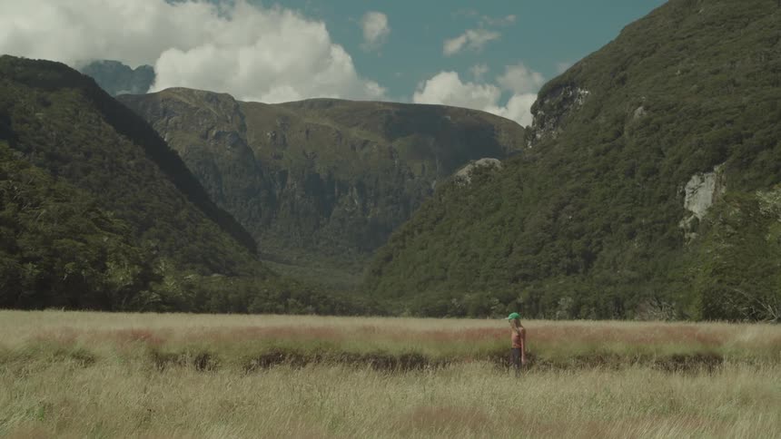 A woman in a green hat walks through a field of dry tall grass with treecovered mountains in the background under a partly cloudy sky