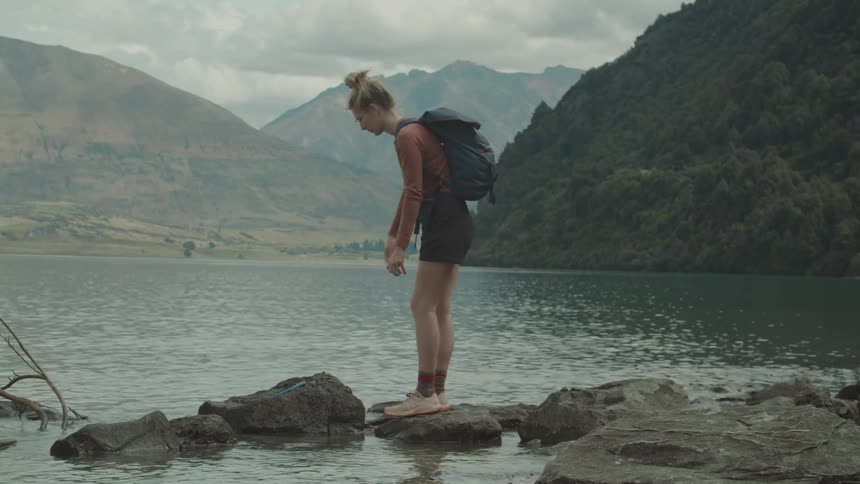 A female hiker squats on lakeside rocks then stands to toss something into the water before walking away framed by distant mountains