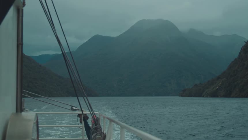 Viewed from the deck of a moving boat a fjord winds between steep treecovered mountains under an overcast sky