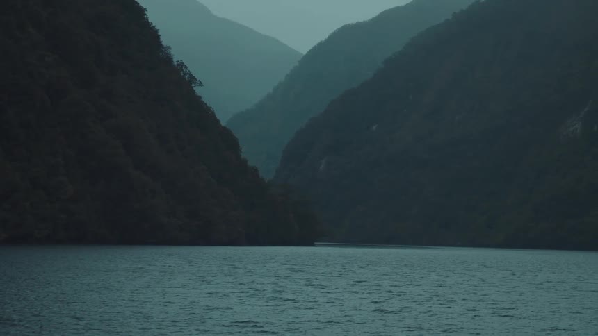 A boat moves slowly across a vast body of water through a moody atmospheric valley surrounded by large silhouetted mountains