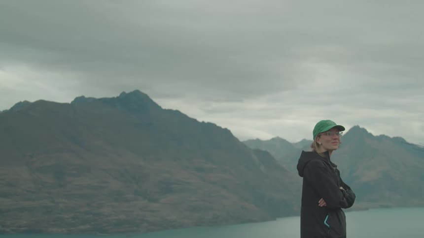A woman wearing a green cap and black jacket stands with her arms crossed looking out at a large lake nestled among mountains under an overcast sky