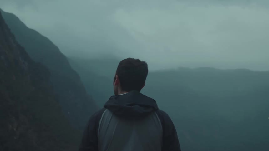 A person wearing a dark jacket with a grey panel stands facing away looking out at a series of dark misty mountains under a cloudy sky