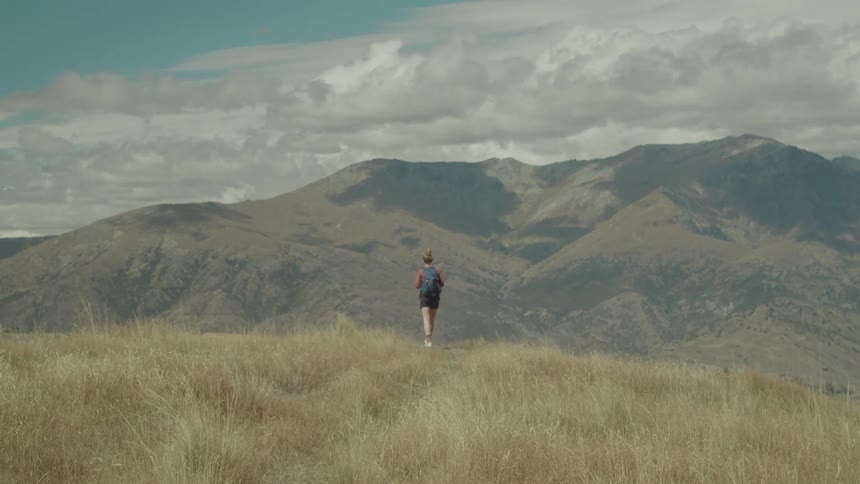 A lone hiker walks in tall grass facing a vast mountain range under a cloudy sky