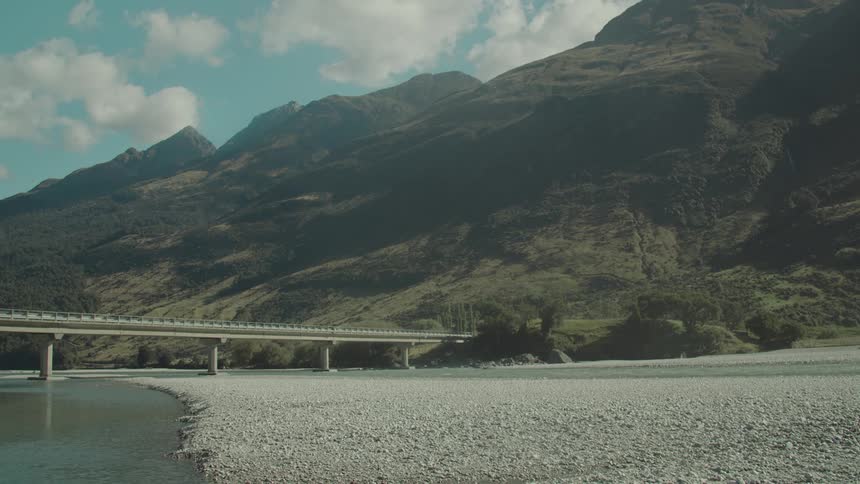 A long bridge stretches over a shallow riverbed beneath a range of sunlit mountains