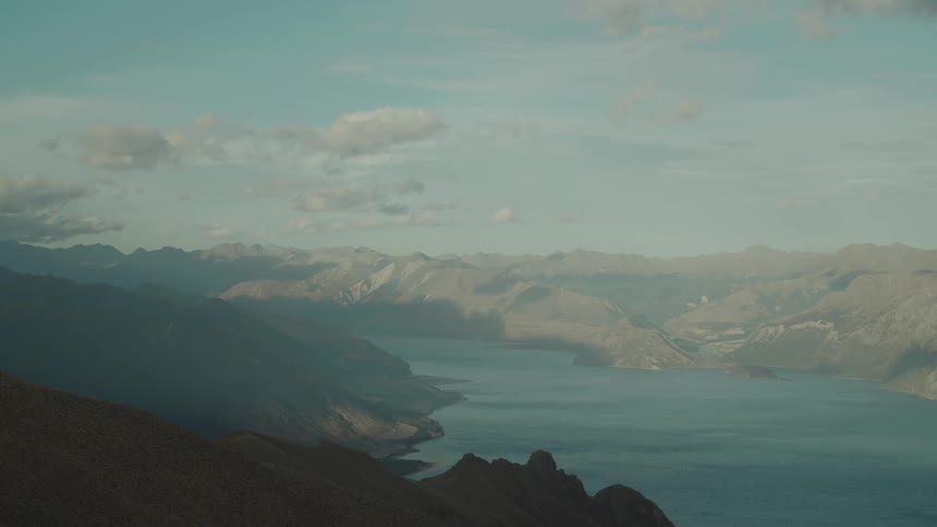 An aerial view shows a large ocean inlet winding through an expansive mountain range under a partly cloudy sky with slopes cast in shadow and light