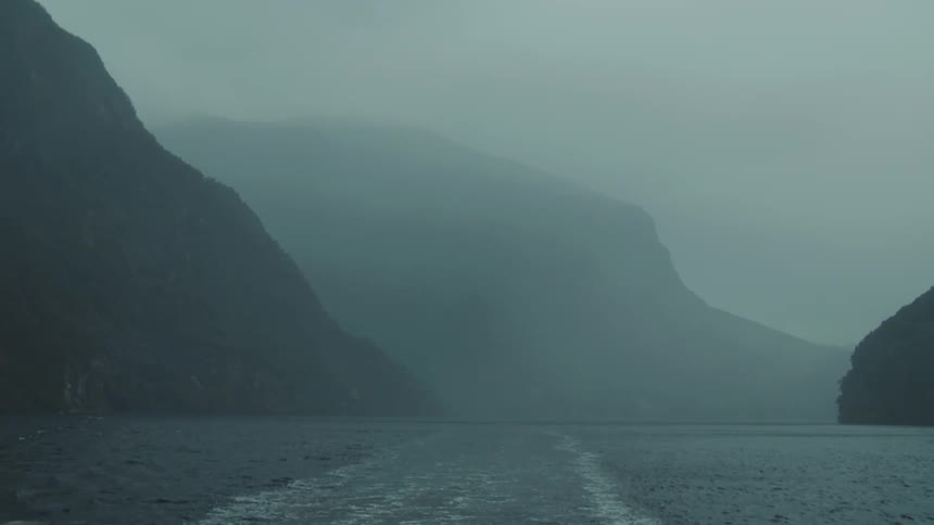 A calm expanse of water extends into the distance flanked by steep dark mountains veiled in light mist with a ships wake trailing behind