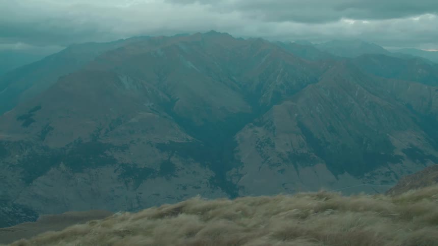 A wide shot captures a vast rugged mountain range under a cloudy sky viewed from a grassy ridge